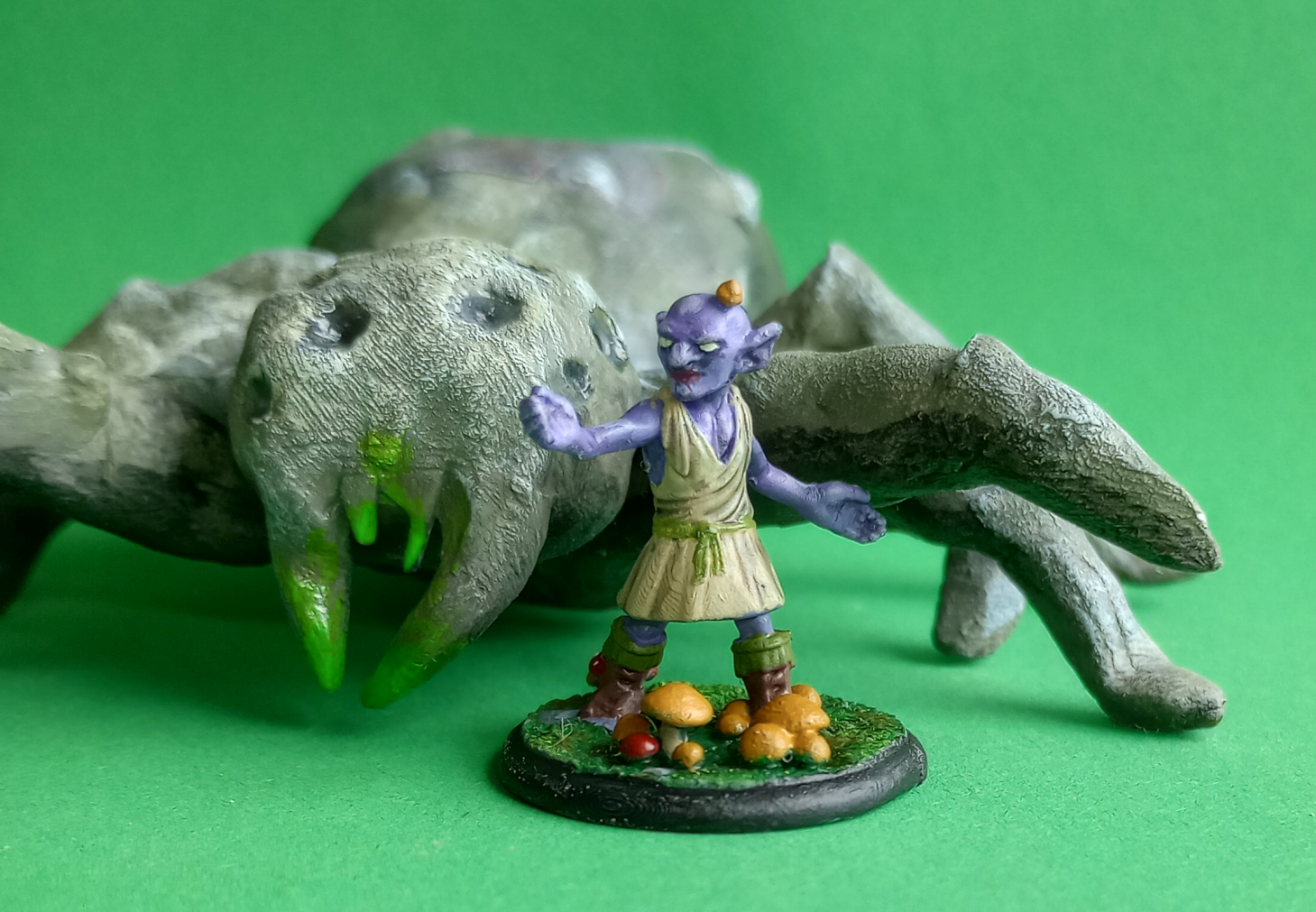 A figurine of a goblin in a field of mushrooms with a mushroom growing out of his head and a giant spider behind him.